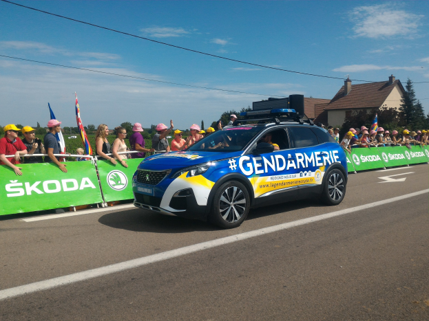 Polizeiauto fährt an einer Menge vorbei, die Fahnen und Mötzen hölt und ein Banner auf dem Auto und Gebäude im Hintergrund, beschriftet mit "Gendarmerie de la Tour de France" unten.