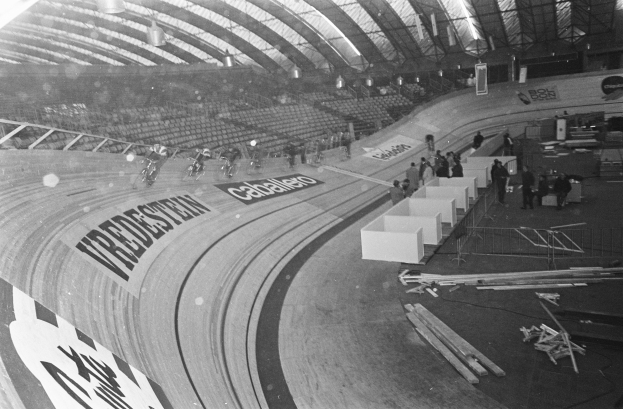 Ein Schwarz-Weiß-Foto eines großen Hallenstadions mit einer Bahn in der Mitte, ein paar Menschen auf der rechten Seite, Stühle auf der linken Seite und ein beleuchtetes Dach oben.