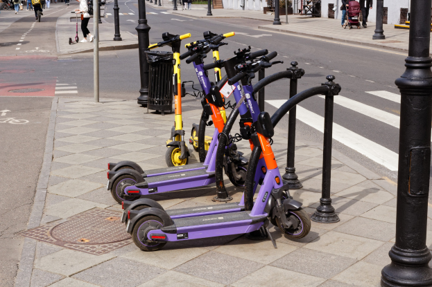 Eine Reihe von E-Scootern, die an einer Straße geparkt sind, mit Fußgängern und Fahrrädern auf dem Gehweg, einem Müllcontainer im Vordergrund und Gebäuden, Bäumen und Pflanzen im Hintergrund.
