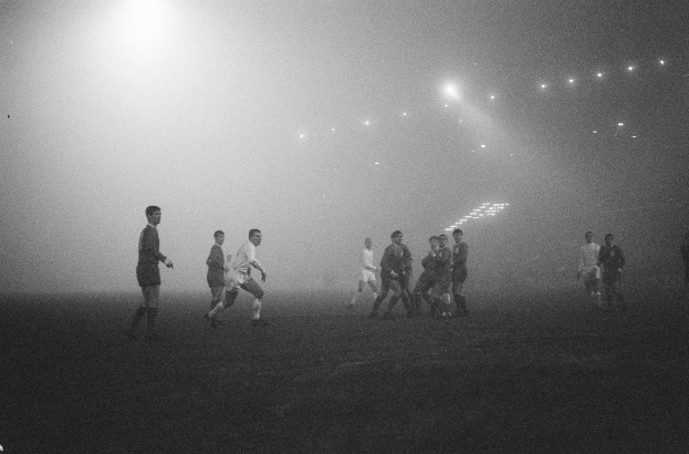 Eine Gruppe von Männern, die Fußball auf einem nebligen Feld spielen, das im Hintergrund von Scheinwerfern beleuchtet wird, in einem Schwarz-Weiß-Bild.