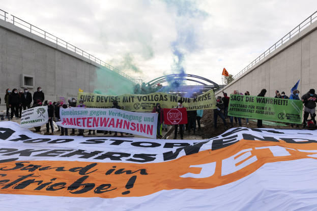 Eine Gruppe von Menschen steht vor einer großen Fahne mit der Aufschrift "Klima-Protest in Berlin", sie halten Schilder und Fahnen in den Händen, im Hintergrund ist Rauch zu sehen und am Himmel eine bewölkte Wolkendecke.