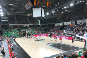 Ein Basketballspiel in einer großen Arena mit Zuschauern auf den Rängen, Anzeigetafeln mit "Asvel vs Galatasaray" und einer Decke mit Lampen und einem Bildschirm.