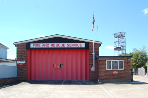 Feuerwehr- und Rettungsdienstgebäude mit roter Tür, Fenstern, einem Namensschild, einem Fahnenmast mit Flagge, einem Metallturm, einem Zaun, Bäumen und einem bewölkten Himmel.