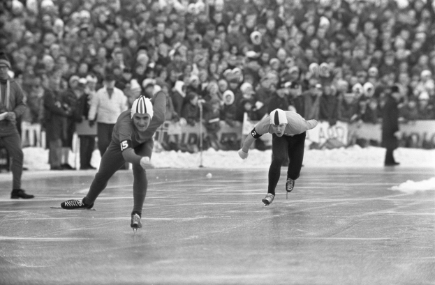 Zwei Männer schnellen auf einer Eisbahn vor einer großen Menge, tragen Helme und Schlittschuhe, mit einem unscharfen Hintergrund in einem Schwarz-Weiß-Bild.