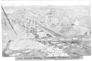 Ein Schwarz-Weiß-Foto eines Kohlebergwerks in Kolumbien, das einige Gebäude, Bäume und Berge im Hintergrund zeigt, mit Text am unteren Bildrand.