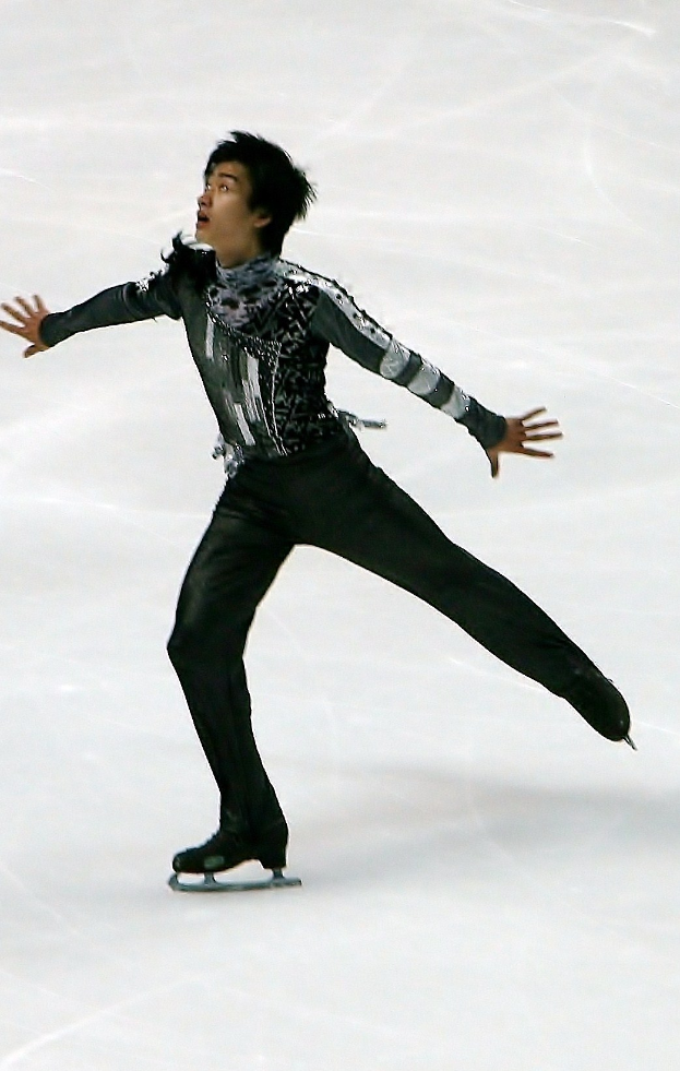 Männlicher Eiskunstläufer in einem schwarzen Kleid, der eine Routine auf einer Eisfläche präsentiert und dabei mit synchronen Arm- und Beinbewegungen gleitet.