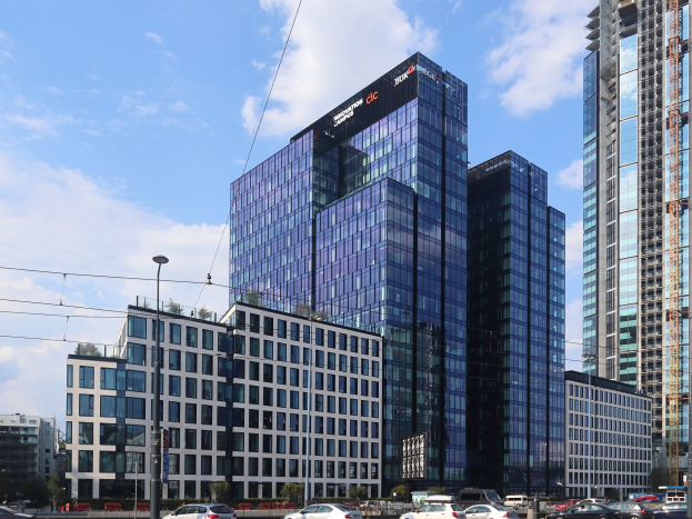 Außenansicht des Deutschen Bank-Hauptsitzgebäudes in Frankfurt, Deutschland, mit umgebender städtischer Infrastruktur einschließlich Straßenlaternen, Fahrzeugen und Bäumen.