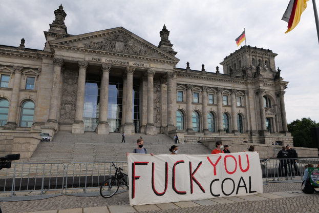 Menschen mit einem 'Fuck You Coal'-Schild vor dem Reichstag in Berlin, mit Bäumen, einer Flagge und einem bewölkten Himmel im Hintergrund.