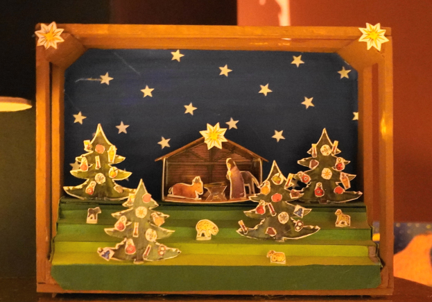 Eine hölzerne Krippe auf einem Tisch mit einer Lampe links und einer dekorierten Kiste mit Weihnachtsbäumen, Tieren und einem Haus im Hintergrund.