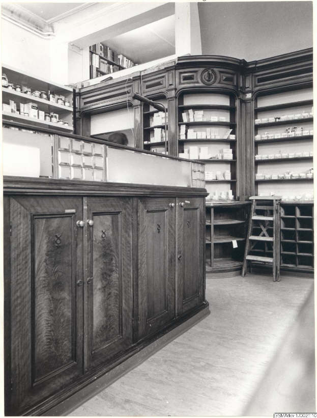 Ein Schwarz-Weiß-Foto eines Apothekeninneren mit einem Tresen, Schränken, einer Leiter und Regalen voller verschiedener Gegenstände, mit Text oben auf dem Bild.