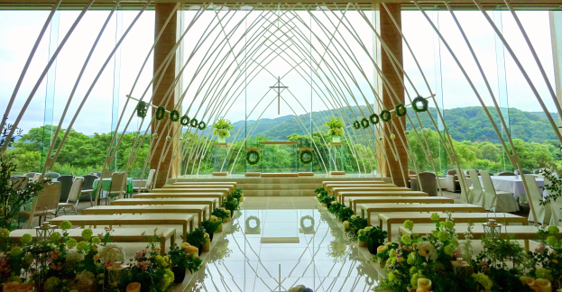 Eine Trauungszeremonie in einem großen Saal mit halbrunder Sitzordnung, Blumenschmuck und einer malerischen Außenansicht von Bäumen, Hügeln und einem blauen Himmel.