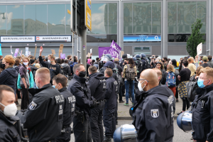 Eine große Gruppe von Menschen vor einem Gebäude stehend, einige mit Schildern und Helmen, mit einem Mast mit einem Schild im Vordergrund und einem Baum im Hintergrund, scheinbar protestierend.