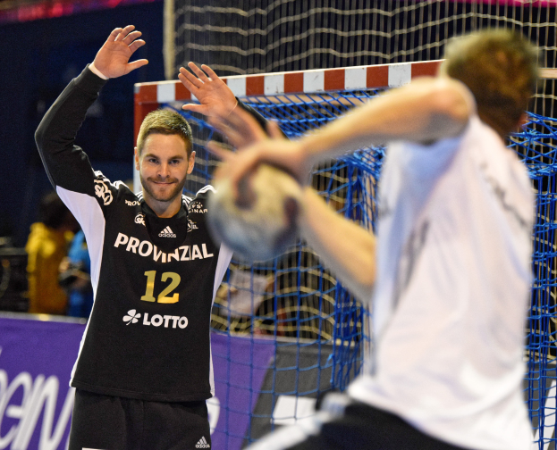Zwei Männer spielen Handball vor einem Tor mit Netz und einer Banner mit Text im Hintergrund und einigen Zuschauern in einem Stadion.