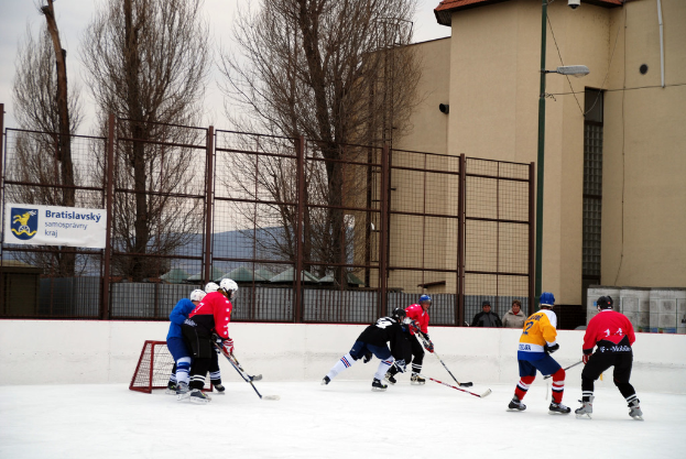 Personen beim Eishockey auf einem Eis mit Gebäuden, Bäumen, einer Straßenlaterne, einem Namensschild und Zäunen im Hintergrund unter einem Himmel.