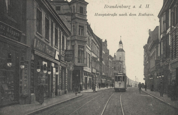 Schwarz-weiß-Foto einer Straße in Brandenburg, Deutschland mit einer Tram, Passanten, Gebäuden, Schildern, Straßeleuchten und einem bewölktem Himmel.