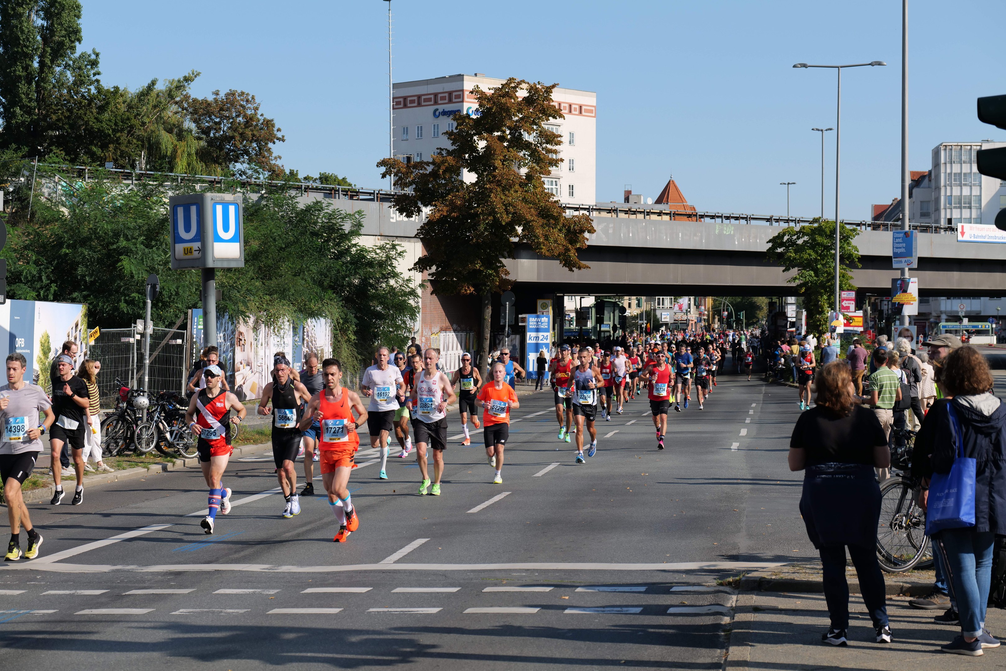 Gruppe von Menschen, die bei einem Marathon auf einer baumgesäumten Straße unter einem klaren blauen Himmel laufen.