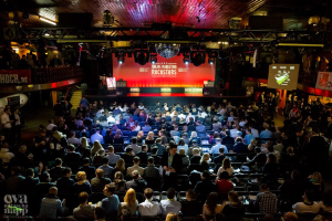 Eine große Menschenmenge sitzt auf Stühlen vor einer Bühne mit Bildschirmen und Plakaten, beleuchtet von Scheinwerfern und umgeben von Absperrungen, voller Vorfreude auf den Beginn eines Online-Marketing-Rockstars-Events, mit einem Wasserzeichen in der linken unteren Ecke.