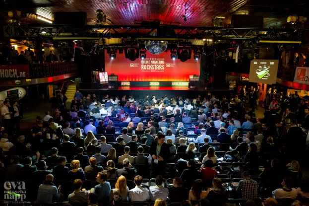 Eine große Menschenmenge sitzt auf Stühlen vor einer Bühne mit Bildschirmen und Plakaten, beleuchtet von Scheinwerfern und umgeben von Absperrungen, voller Vorfreude auf den Beginn eines Online-Marketing-Rockstars-Events, mit einem Wasserzeichen in der linken unteren Ecke.