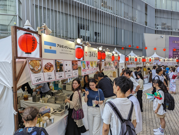 Menschen um einen Tisch mit Essen auf einem Outdoor-Event, einige tragen Taschen und halten Gl├Ąser, mit Informationsplakaten und h├Ąngenden Laternen über ihnen, vor einem Gebäudebackgrund in Japan.