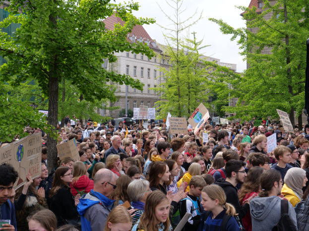 Eine große Menschenmenge mit Schildern versammelt sich vor einem Gebäude in Berlin, mit Bäumen, Fahrzeugen und einem Redner im Hintergrund.