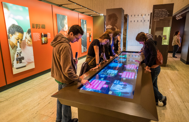 Eine Gruppe von Menschen bei einer Museumsführung sitzt an einem Tisch und betrachtet einen Bildschirm, mit Fotorahmen an der linken Wand und einer Tafel mit Text an der rechten Seite.