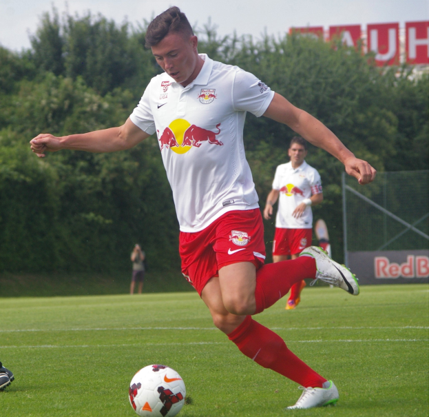 Ein Mann in einem Fußballtrikot kickt einen Ball auf einem grünen Rasen mit Bäumen und einem klaren blauen Himmel im Hintergrund, mit einem Schild mit der Aufschrift "RB Leipzig" im Bild.