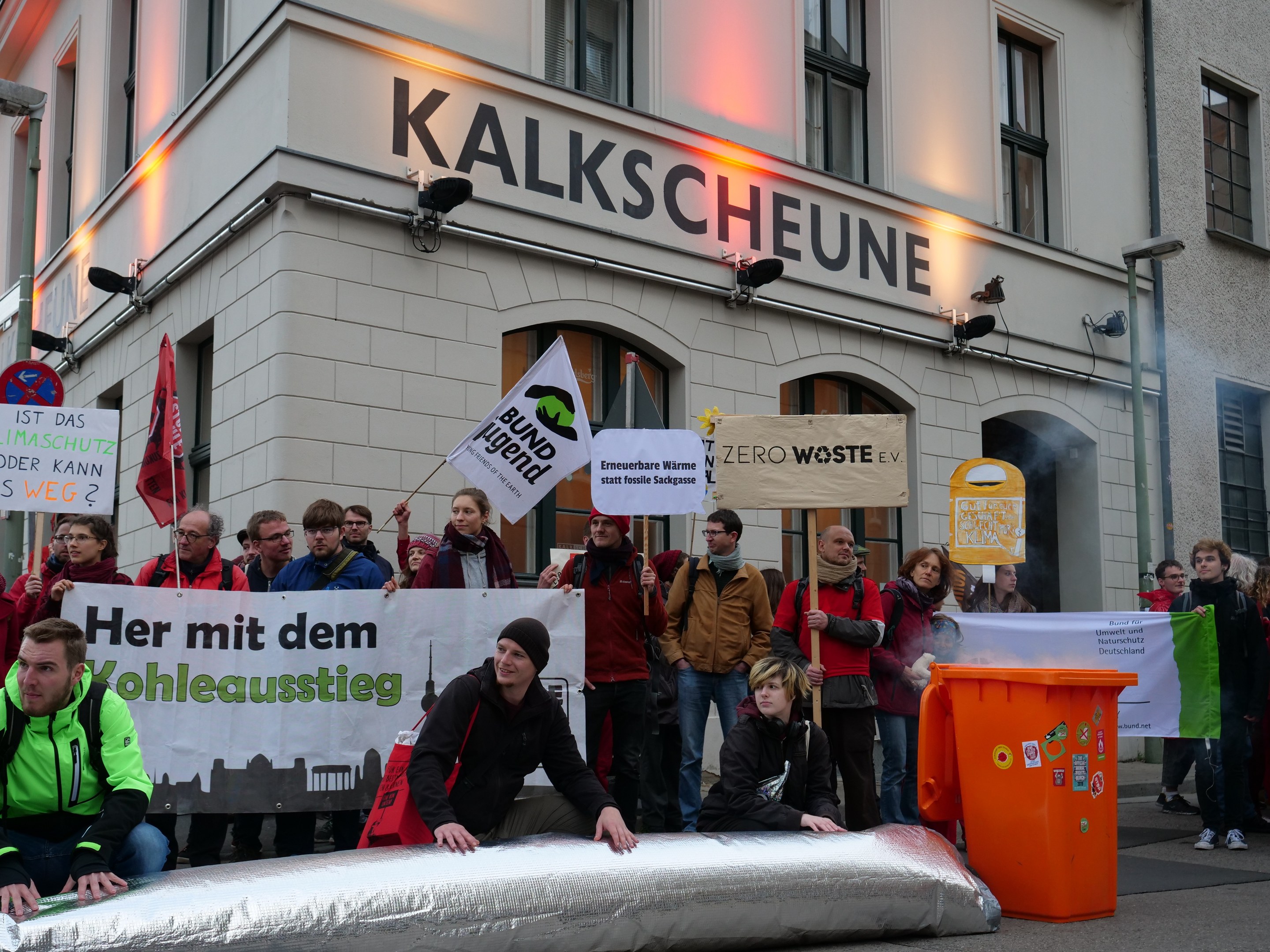 Eine Gruppe von Menschen mit Protestschildern und Plakaten vor einem Gebäude in Deutschland, mit zwei Personen im Vordergrund und einem Müllcontainer rechts.