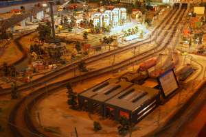 Eine detaillierte Modellbahnausstellung in einem Museum, die Gebäude, Bäume, Eisenbahnschienen, Fahrzeuge, Masten, Lichter und Platten zeigt, die eine belebte Stadtlandschaft nachahmen.