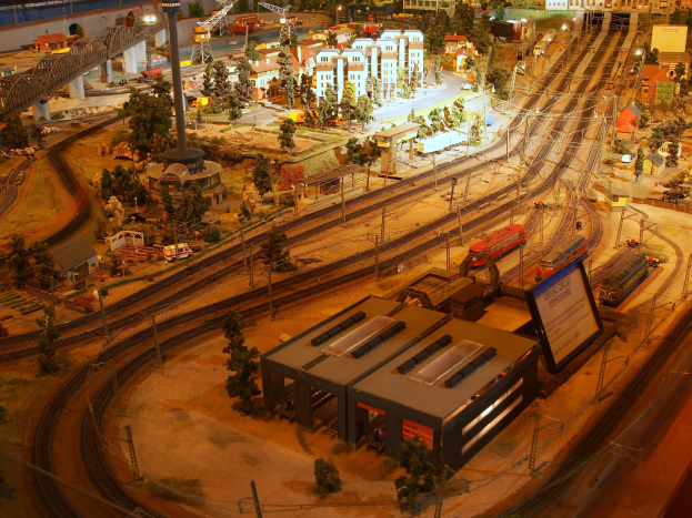 Eine detaillierte Modellbahnausstellung in einem Museum, die Gebäude, Bäume, Eisenbahnschienen, Fahrzeuge, Masten, Lichter und Platten zeigt, die eine belebte Stadtlandschaft nachahmen.