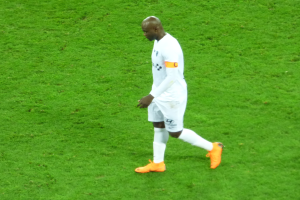 Ein Fußballspieler in einem weißen T-Shirt, weißen Shorts und orangefarbenen Schuhen läuft über ein saftiges grünes Feld.
