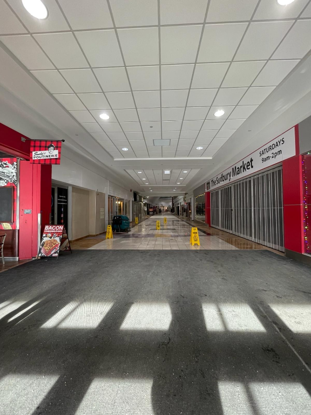 Innenansicht eines Einkaufszentrums mit Deckenbeleuchtung, einem Stuhl und einer Tafel mit der Aufschrift "Sydbury Market - Vorher und Nachher" links und verstreuten Gegenständen rechts und auf dem Boden.