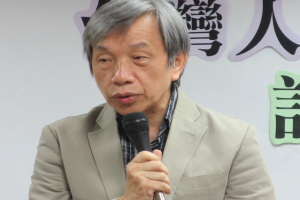 Ein Mann im Anzug spricht vor einer textbedeckten Wand in ein Mikrofon und sieht ernst und professionell aus.