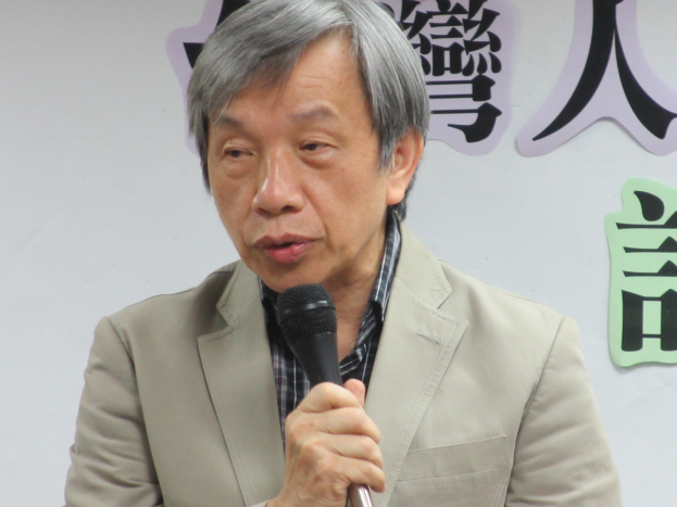 Ein Mann im Anzug spricht vor einer textbedeckten Wand in ein Mikrofon und sieht ernst und professionell aus.