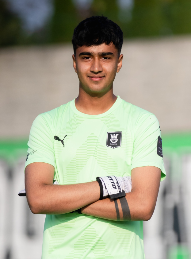 Ein junger Mann in einem grünen T-Shirt steht auf einem Fußballfeld und hat die Arme verschränkt, er lächelt und der Hintergrund ist unscharf.