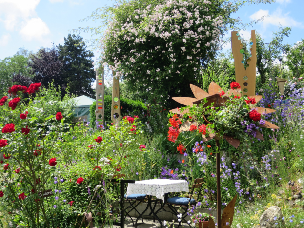 Ein farbenfroher Garten auf dem Chelsea Flower Show mit bunten Blumen und Pflanzen, einem zentralen Tisch mit einer Decke und Stühlen, umgeben von Bäumen unter einem bewölkten Himmel.