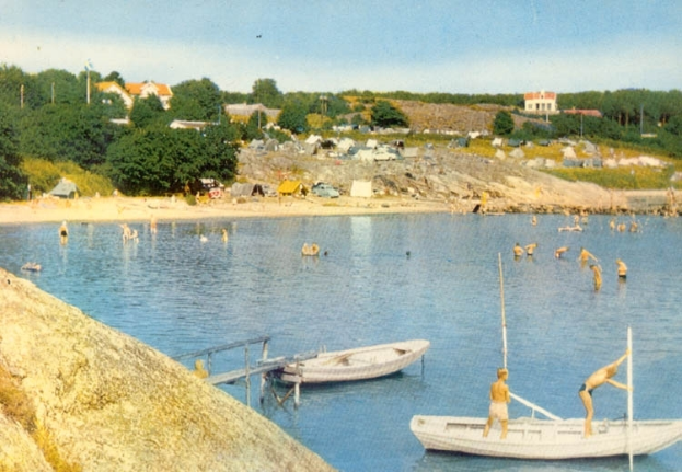Ein altes Foto von Menschen, die in der Nähe eines Bootes schwimmen, mit einer Brücke und Geländern im Vordergrund, einem Felsen links und Bäumen, Gebäuden, Fahrzeugen, Masten und einem bewölktem Himmel im Hintergrund.