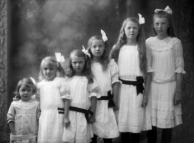 Schwarz-weiß-Fotografie einer Gruppe junger Mädchen in weißen Kleidern, die in einer Reihe vor einer Wand stehen.