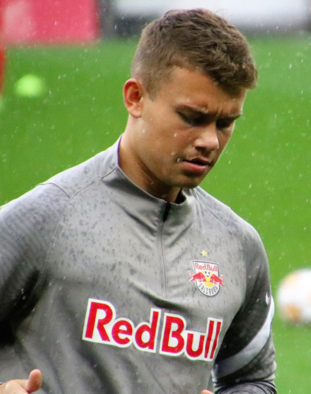 Ein Mann in grauem Hemd steht auf einem Fußballfeld mit ausgestreckten Armen und entschlossener Miene während eines Trainings, mit einem unscharfen Hintergrund.