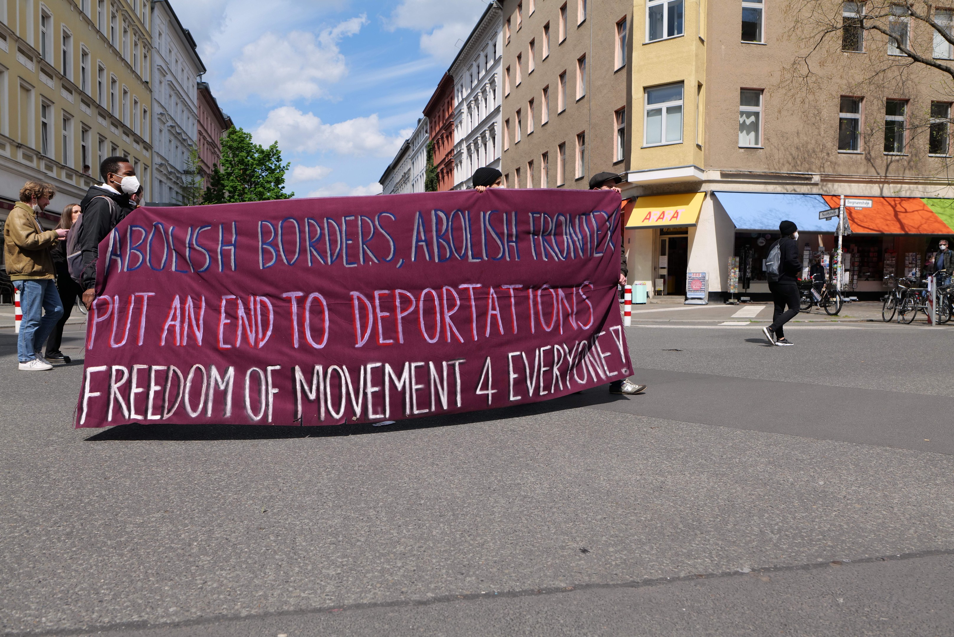 Eine Gruppe von Menschen marschiert auf einer Stadtstraße und hält ein Transparent mit der Aufschrift "Abolish Borders, Abolish Frontiers, Put an End to Deportations, Freedom of Movement 4 Everyone" hoch, mit Gebäuden, Bäumen, Fahrrädern und bewölktem Himmel im Hintergrund.