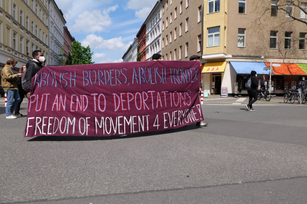 Eine Gruppe von Menschen marschiert auf einer Stadtstraße und hält ein Transparent mit der Aufschrift "Abolish Borders, Abolish Frontiers, Put an End to Deportations, Freedom of Movement 4 Everyone" hoch, mit Gebäuden, Bäumen, Fahrrädern und bewölktem Himmel im Hintergrund.