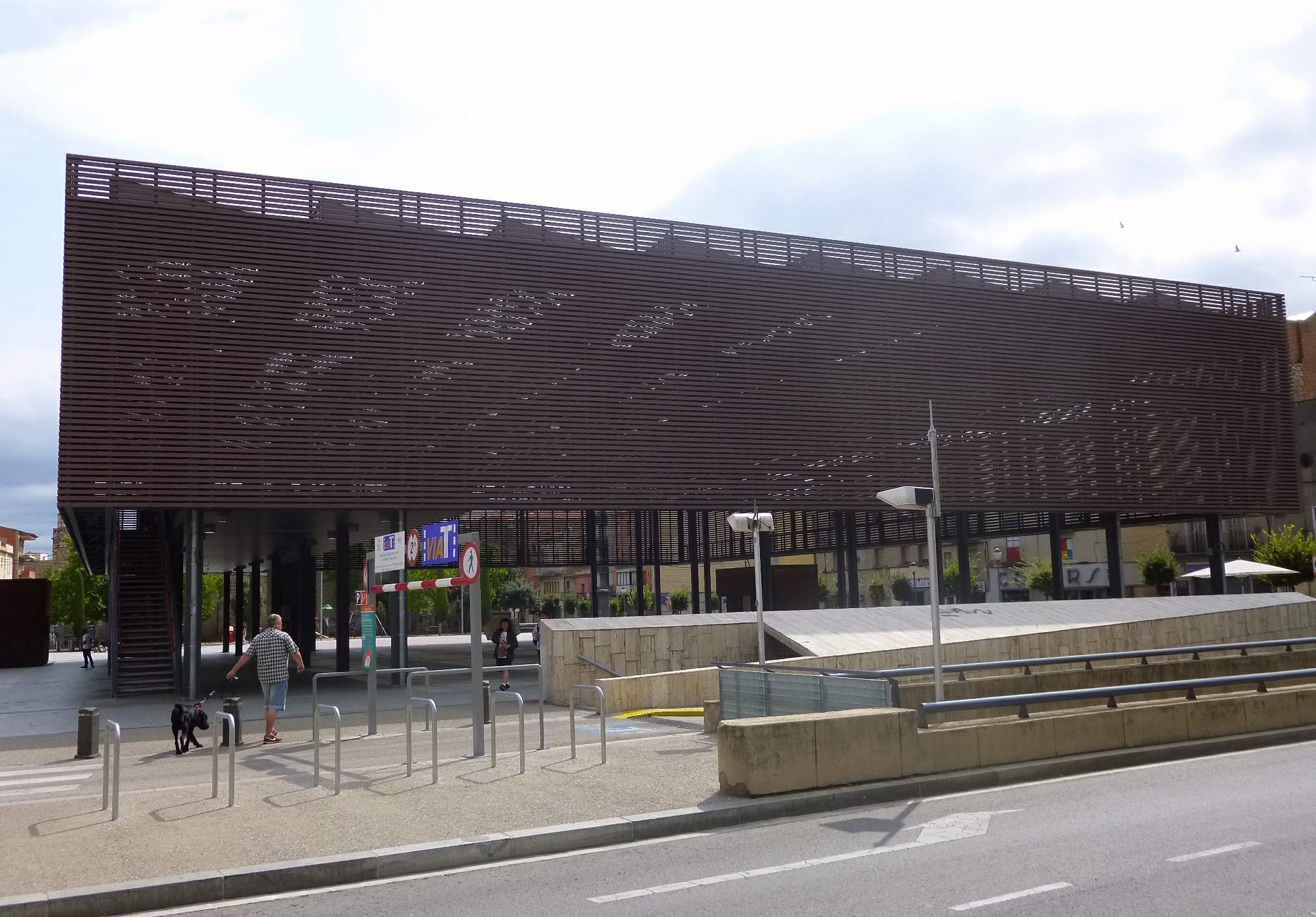 Großes modernes Gebäude mit zahlreichen Fenstern, das als neuer Eingang zur Stadt dient, umgeben von städtischer Infrastruktur einschließlich Straßenlaternen, Fahrzeugen, Fußgängern und Bäumen unter einem bewölkten Himmel.