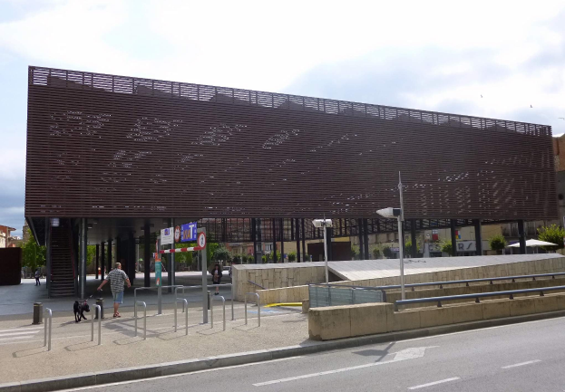 Großes modernes Gebäude mit zahlreichen Fenstern, das als neuer Eingang zur Stadt dient, umgeben von städtischer Infrastruktur einschließlich Straßenlaternen, Fahrzeugen, Fußgängern und Bäumen unter einem bewölkten Himmel.