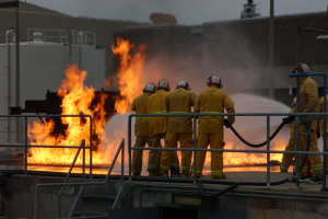 Feuerwehrleute in Helmen stehen auf einem Dach und halten Schläuche, mit Geländern und Treppen darunter und einem Gebäude und Himmel im Hintergrund.