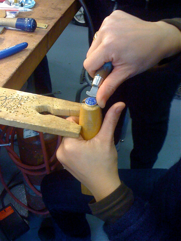 Ein Mann graviert Holz mit einem Meißel an einem Tisch mit Werkzeugen, während ein anderer Mann daneben steht.