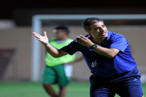 Ein Mann in einem blauen T-Shirt und Hose steht auf einem Fußballfeld und hält ein Mikrofon in der Hand, während er seinen Spielern Zeichen gibt. Im Hintergrund ist eine Person in einem grünen Kleid, ein Torpfosten und eine Wand zu sehen.