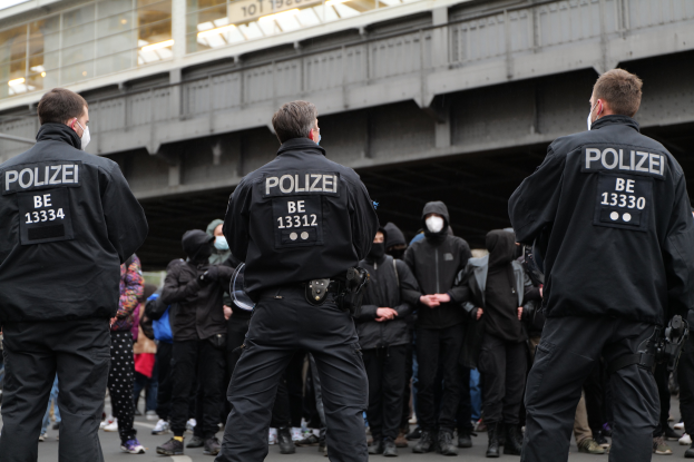 Eine Gruppe von Polizisten in Uniform steht vor einer Menge von Menschen in schwarzen Uniformen und Masken mit einer Brücke und einem Gebäude im Hintergrund während einer Protestaktion in einer Stadt.