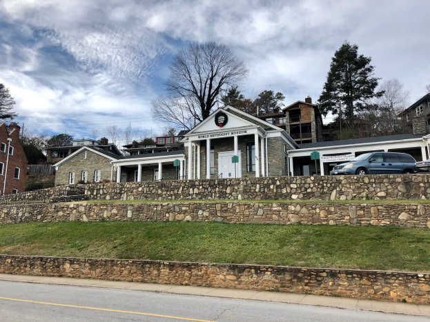 Steinmauer mit parkenden Autos davor, umgeben von Gebäuden, Bäumen und einem bewölkten Himmel, als Weltkulturerbe-Museum bezeichnet.