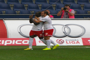 Zwei Fussballspieler feiern ein Tor auf einem Feld, umgeben von einer Gruppe von Menschen, mit einer Person, die eine Kamera hält, einem Banner mit Text, Stühlen und einer Wand im Hintergrund.
