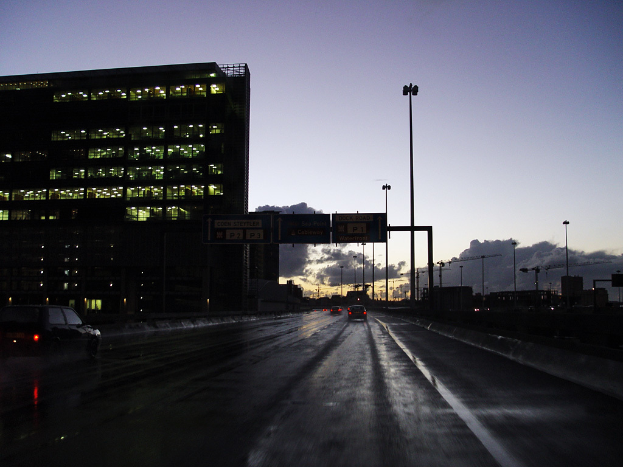 Ein transparentes Gebäude voller Lichter, mit mehreren Strommasten und einer Straße mit ein paar Fahrzeugen auf der rechten Seite.