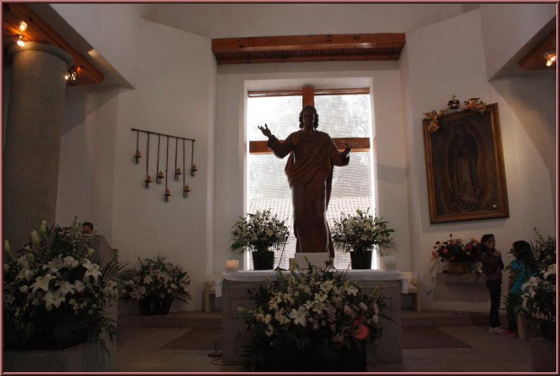 Ein Innenblick in ein Gebäude mit Kindern auf dem Boden, einer Skulptur, einer mit Stoff bedeckten Wand, Topfpflanzen, einem Fenster, einer Säule mit einem Holzfuß und Lampen sowie einem an der Wand befestigten Photo-Rahmen.